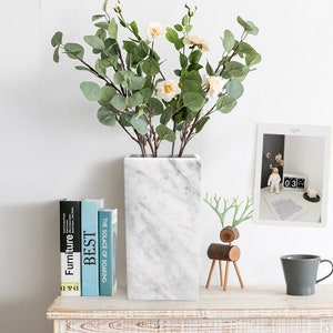 Op de afbeelding: Witte marmeren vaas met een vierkante vorm en een boeket witte bloemen en groene bladeren. De vaas staat op een houten tafel met boeken, een houten hertenbeeldje en een kopje.