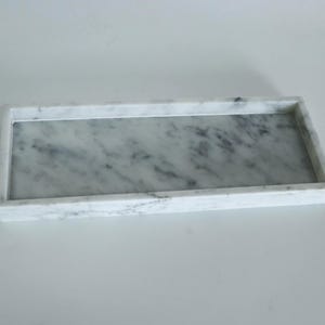 Puede incluir: Una bandeja rectangular de mármol blanco con una superficie lisa. La bandeja tiene un borde elevado alrededor del perímetro.