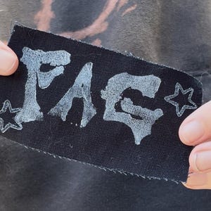 May include: A rectangular black fabric patch featuring the white hand-painted text FAQ in a stylized, distressed font. The design is flanked by a small white five-pointed star on both the left and right sides, with raw, unfinished edges.