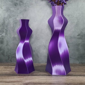 May include: Two purple decorative vases with a unique, twisted design. The larger vase holds a bundle of purple decorative branches. The vases are made of a shiny material and sit on a wooden surface.