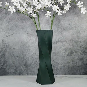 May include: A dark green, geometric vase holds white flowering branches. The vase has a twisted design and sits on a white surface against a gray textured background. The flowers have small, delicate white petals.