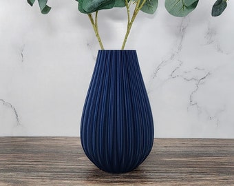 May include: A navy blue vase with vertical ridged texture holds faux eucalyptus branches. The leaves are a muted green, and the vase sits on a wooden surface against a marble background. A decorative floral arrangement.