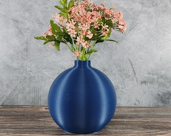 May include: A navy blue, spherical vase holds a bouquet of pink flowers with green leaves. The vase sits on a wooden surface against a gray textured background. The vase has a smooth, matte finish.