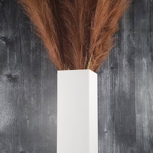 May include: A white rectangular vase holds a bundle of dried, feathery brown pampas grass. The vase is set against a dark gray wooden background. The arrangement is simple and elegant, suitable for home decor.