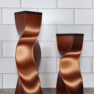 May include: Two copper-colored, twisted decorative vases with a textured surface. The vases have a square base and top with a black border. The background is a white tiled wall. The vases are of different sizes.