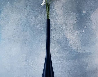 May include: A tall, navy blue vase with a slender neck and flared base, holding white flowering branches. The vase is set against a textured, blue-gray backdrop, creating a minimalist aesthetic. The vase is approximately 12 inches tall.