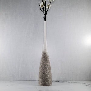 May include: A tall, slender vase with a white top that fades into a textured, speckled brown base. The vase holds a branch with small white flowers. The vase is a decorative home accent.