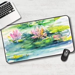 May include: Watercolor mousepad featuring a serene pond scene with pink and yellow water lilies and green lily pads. The design has a loose, artistic style with black outlines and a blue and green water background. A black computer mouse is next to the mousepad.