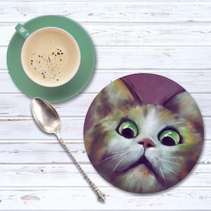 May include: A round cat coaster with a painted image of a cat's face with large green eyes. A green teacup and saucer with coffee and a silver spoon are next to the coaster on a white wooden surface.
