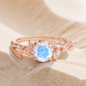 Könnte beinhalten: Roségoldener Ring mit einem großen, runden, schillernden Edelstein. Das Band ist mit filigranen Blatt- und Rankendetails verziert. Der Ring ist vor einem sandigen Hintergrund platziert, der sein elegantes Design hervorhebt. Ein wunderschönes Schmuckstück.