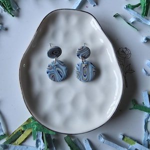 May include: A pair of handmade polymer clay earrings. Each earring features a circular design with blue, tan, and navy swirls. The earrings are displayed on a white, irregularly shaped plate with a gold rim.