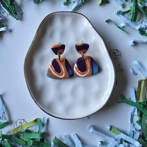 May include: A pair of colorful polymer clay earrings with a marbled design. The earrings feature a dark purple base with orange, white, and blue swirls. They are displayed on a white, organically shaped plate with a gold rim, surrounded by colorful paper shreds.