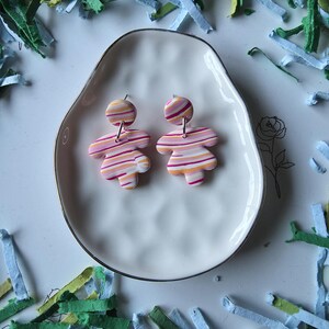 May include: A pair of handmade polymer clay earrings. Each earring features a striped design in shades of pink, orange, and white. The earrings are on a white, irregularly shaped plate with a silver rim. The background includes colorful paper shreds.