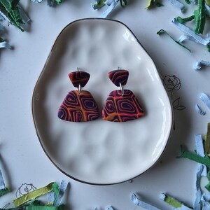 May include: A pair of handmade polymer clay earrings. The earrings feature a geometric design with shades of purple, orange, and red. They are displayed on a white, irregularly shaped plate with a gold rim. The background includes colorful paper scraps.