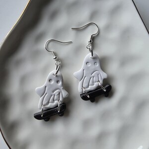 May include: A pair of Halloween-themed earrings. Each earring features a white ghost figure riding a black skateboard. The earrings have silver-colored hooks. The earrings are displayed on a white plate with a gold rim.