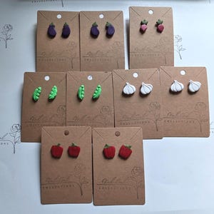 May include: A collection of nine pairs of stud earrings shaped like miniature vegetables, including purple eggplants, pink radishes, green pea pods, white garlic bulbs, and red bell peppers, each mounted on individual brown cardboard display cards.