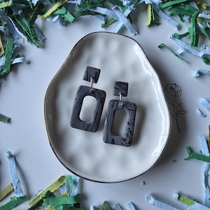 May include: A pair of gray marbled polymer clay earrings with a rectangular shape, displayed on a white, irregularly shaped plate with a silver rim. The earrings have a square top and a rectangular drop. The background features colorful paper confetti.