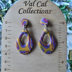 May include: A pair of teardrop-shaped earrings with a swirl pattern of yellow, purple, and blue. The earrings are hanging on a wooden display board with the text "Val Cal Collections". The board has a ruler marked in inches.