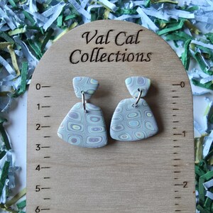 May include: A pair of handmade polymer clay earrings with a geometric pattern in shades of blue, purple, and green. The earrings are displayed on a wooden measuring board with the text "Val Cal Collections".