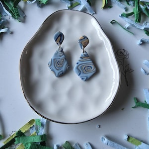 May include: A pair of handmade, marbled polymer clay earrings. Each earring features a diamond shape with blue, tan, and navy swirls. The earrings hang from silver-colored hooks. They are displayed on a white, scalloped plate with a gold rim, surrounded by colorful paper.