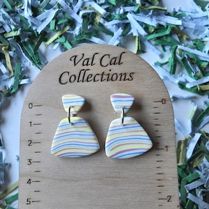 May include: A pair of colorful, striped earrings with a geometric design. The earrings are displayed on a wooden measuring board with the text "Val Cal Collections". The earrings have a yellow, blue, and pink color scheme.