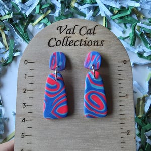 May include: A pair of colorful earrings with a blue, red, and white swirled pattern. The earrings are displayed on a wooden stand with the text "Val Cal Collections". The stand has a ruler printed on it.