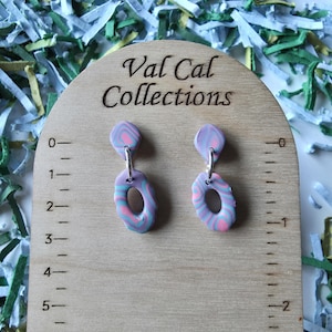 May include: A pair of dangle earrings with a marbled design in shades of lavender, pink, and teal. The earrings are connected by silver-colored metal hoops. The wooden display stand has the text "Val Cal Collections".