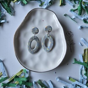 May include: A pair of gray oval dangle earrings with a speckled pattern, displayed on a white, organically shaped dish. The earrings have a circular top and a larger oval drop. The background includes colorful paper shreds.