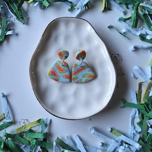 May include: A pair of colorful polymer clay earrings with a unique abstract design. The earrings feature a light blue base with orange, pink, and green swirls. They are displayed on a white, irregularly shaped plate with a silver rim, surrounded by colorful paper shreds.
