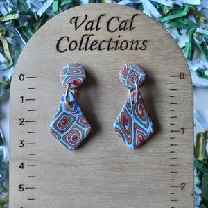 May include: A pair of colorful, patterned earrings with a diamond shape, displayed on a wooden stand. The earrings feature a swirl design in shades of blue, red, orange, and white. The wooden stand has the text "Val Cal Collections".