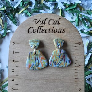 May include: A pair of colorful, abstract polymer clay earrings with a marbled design in shades of orange, blue, yellow, and red. The earrings are displayed on a wooden measuring board with the text "Val Cal Collections".