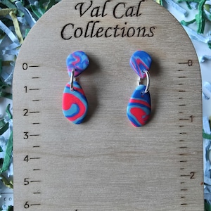 May include: A pair of handmade earrings with a marbled design in red, blue, and white. The earrings are teardrop-shaped and hang from silver-colored hoops. The wooden display stand has the text "Val Cal Collections".