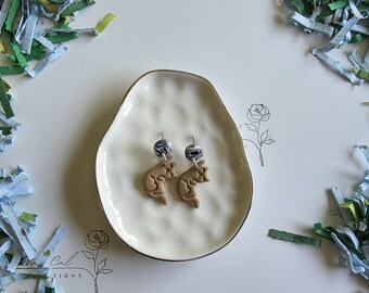 May include: A pair of handmade earrings with a light brown kangaroo design. The earrings are displayed on a white, organic-shaped dish with a gold rim. The earrings have a marbled bead at the top. The background includes paper confetti.