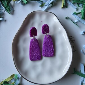 May include: A pair of purple earrings with a floral pattern, displayed on a white, oval-shaped plate. The earrings have a rectangular drop with a smaller, rounded top. The plate is surrounded by colorful paper strips.
