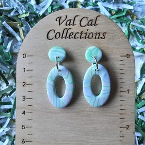 May include: A pair of oval-shaped earrings with a marbled design in shades of green, blue, and pink. The earrings are suspended from a small, round stud. The earrings are displayed on a wooden surface with the text "Val Cal Collections".