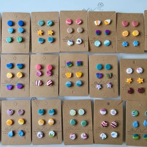 May include: A collection of handmade stud earrings displayed on brown cards. The earrings feature various shapes and colors, including hearts, stars, and circles, with designs in blue, pink, yellow, and green. The earrings are arranged in sets of three.