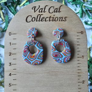 May include: A pair of colorful, abstract earrings with a light blue base and swirling patterns of red, yellow, and orange. The earrings are displayed on a wooden measuring board with the text "Val Cal Collections".