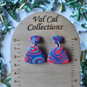 May include: A pair of colorful earrings with a blue, red, and white swirled pattern. The earrings are hanging from silver hooks. The text "Val Cal Collections" is visible on the wooden display board. The earrings are approximately 1 inch in length.