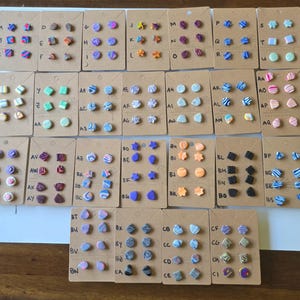 May include: A collection of numerous pairs of small stud earrings displayed on individual brown cardboard cards, each labeled with a letter. The earrings feature a wide variety of shapes, including stars, circles, squares, and abstract forms, in many colors.