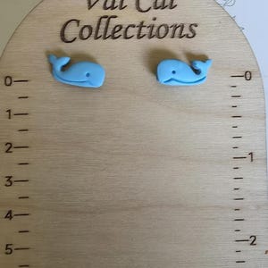 Könnte beinhalten: Ein Paar hellblaue, walformige Ohrstecker auf einer hellen Holzkarte mit dem Text Val Cal Collections. Die Karte ist mit einem aufgedruckten Lineal versehen, das auf der linken Seite Zoll und auf der rechten Seite Zentimeter anzeigt.