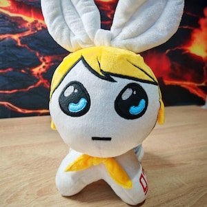 Może przedstawiać: Pluszowa zabawka w kształcie królika z białym korpusem, dużymi białymi uszami i żółtą głową z blond włosami. Zabawka ma duże czarne oczy z niebieskimi akcentami i żółtą chustą. Zabawka stoi na drewnianej powierzchni.