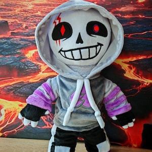 Op de afbeelding: Een pluche speelgoed van een personage met een schedelachtig gezicht, gekleed in een grijze hoodie en paarse en zwarte kleding. Het personage heeft rode ogen en een brede grijns. De achtergrond is een vurige, lava-achtige scène.