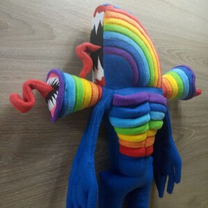 Puede incluir: Un juguete de peluche de color arco&iacute;ris con una boca grande y dentada y brazos y piernas largos y azules. El juguete est&aacute; hecho de tela suave y peluda y tiene un aspecto caprichoso y de dibujos animados.