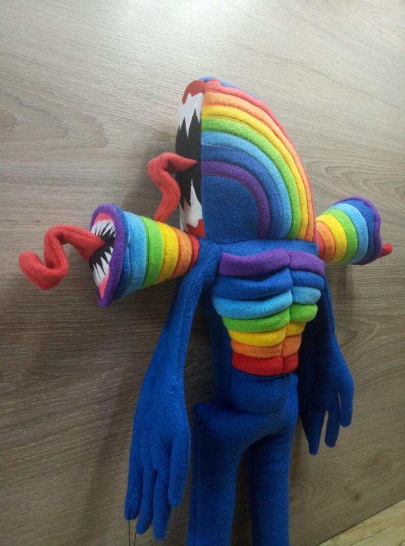 Puede incluir: Un juguete de peluche de color arco&iacute;ris con una boca grande y dentada y brazos y piernas largos y azules. El juguete est&aacute; hecho de tela suave y peluda y tiene un aspecto caprichoso y de dibujos animados.
