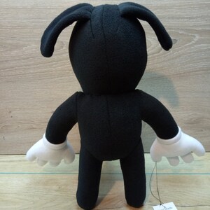 May include: Black plush toy with white gloves and long ears. The toy is standing on a wooden surface. A tag attached to the toy reads 'Hand Made 100% Polyester'. The tag also has symbols for washing instructions.