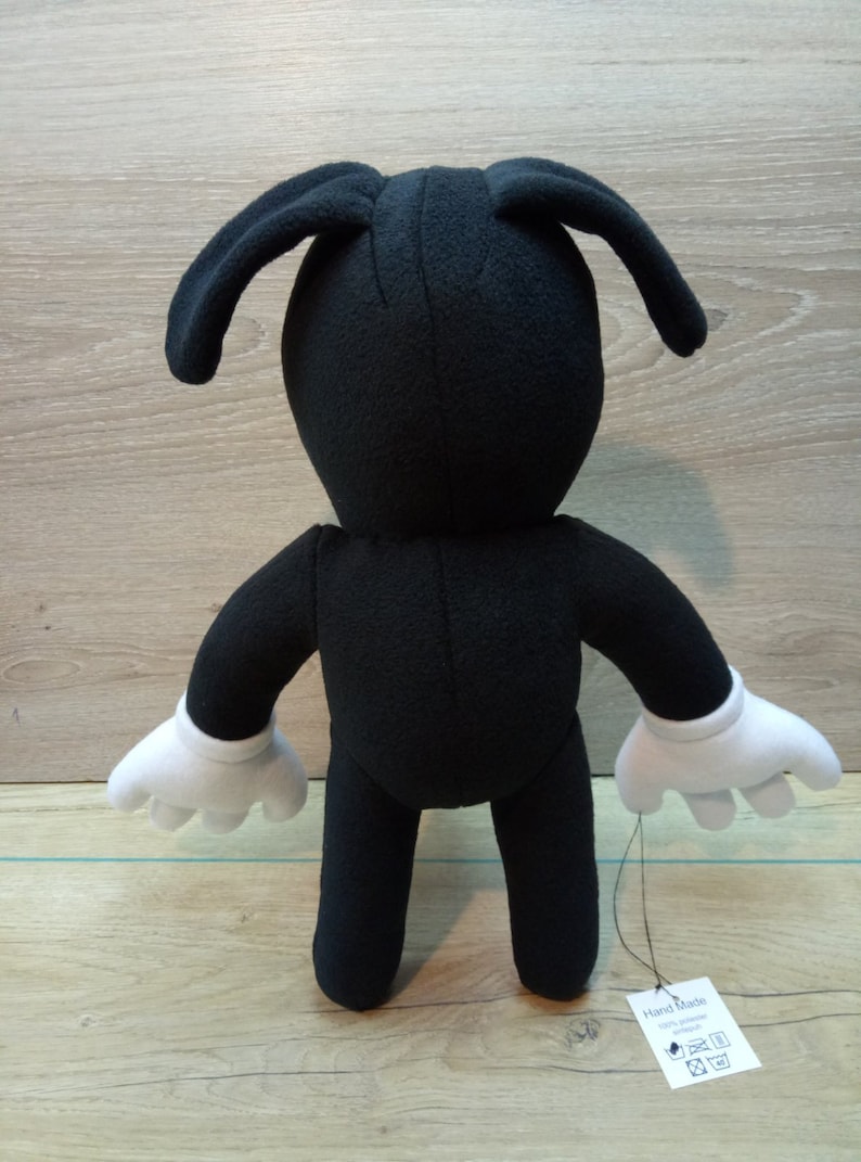 May include: Black plush toy with white gloves and long ears. The toy is standing on a wooden surface. A tag attached to the toy reads 'Hand Made 100% Polyester'. The tag also has symbols for washing instructions.