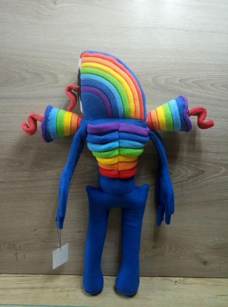 Puede incluir: Un juguete de peluche azul con rayas de arco&iacute;ris y un cuerpo largo y delgado. El juguete tiene dos brazos largos y dos piernas largas. El juguete est&aacute; de pie sobre una superficie de madera.