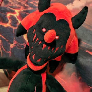 May include: A plush toy featuring a red and black colour scheme with a pointed head, two black horns, and a wide, grinning mouth filled with sharp teeth. The face has two small white eyes, and the torso displays a black centre with a red star emblem.