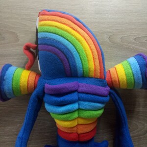 Puede incluir: Un juguete de peluche con forma de criatura arco&iacute;ris con extremidades azules y un torso de color arco&iacute;ris. El juguete est&aacute; hecho de tela suave y peluda.