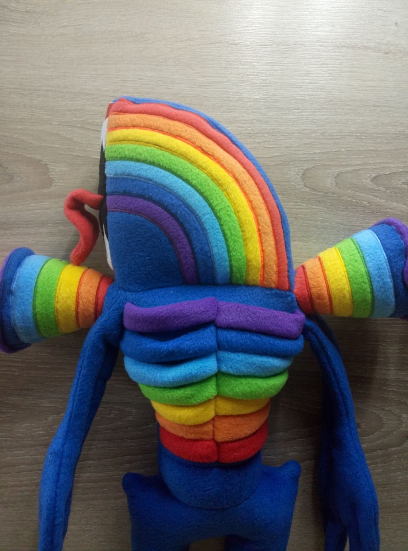 Puede incluir: Un juguete de peluche con forma de criatura arco&iacute;ris con extremidades azules y un torso de color arco&iacute;ris. El juguete est&aacute; hecho de tela suave y peluda.